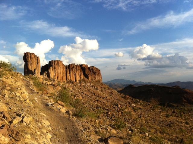 A rugged trail winding through a rocky landscape, leading towards towering rock formations under a blue sky dotted with white clouds. The scene captures a blend of earthy tones and natural textures, showcasing the beauty of the outdoors. Monolith Gardens mountain bike trail.