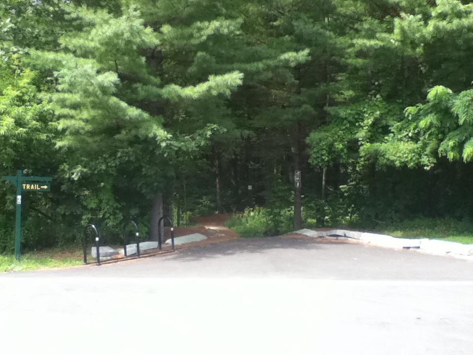 A pathway leading into a wooded area, marked by a sign indicating "TRAIL." In the foreground, there are bike racks, and the surroundings are filled with lush green trees, suggesting a natural and serene setting for outdoor activities. The ground is gravelly, and the trail appears well-defined. Pine Ridge mountain bike trail.