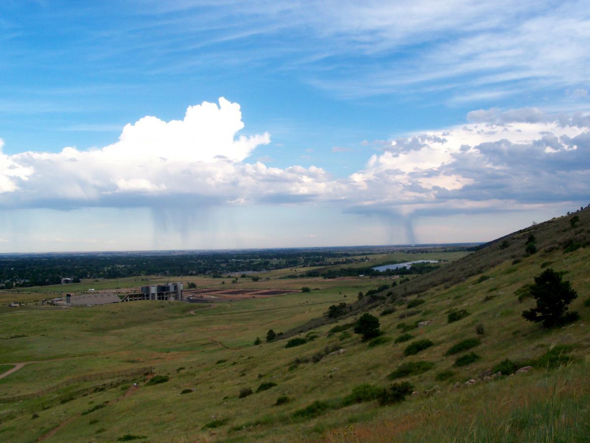 A panoramic view of a lush green landscape under a partly cloudy sky, featuring distant rain showers visible in the background. The foreground includes rolling hills, scattered trees, and a structure on a flat area, with a body of water seen to the right. Foothills Trail mountain bike trail.