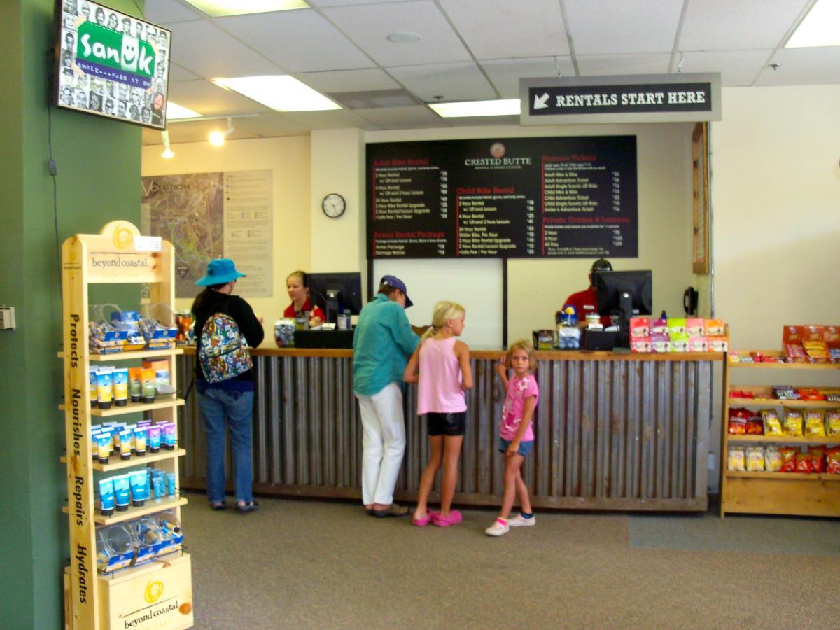 Interior of a rental shop featuring a service desk where customers are purchasing items. Two children are standing near the desk, while an adult wearing a blue hat is interacting with the staff. A display rack is visible on the left with various sunscreen products, and shelves on the right contain snacks. A sign above the desk indicates where rentals begin. Evolution Bike Park at Crested Butte Mountain Resort mountain bike trail.