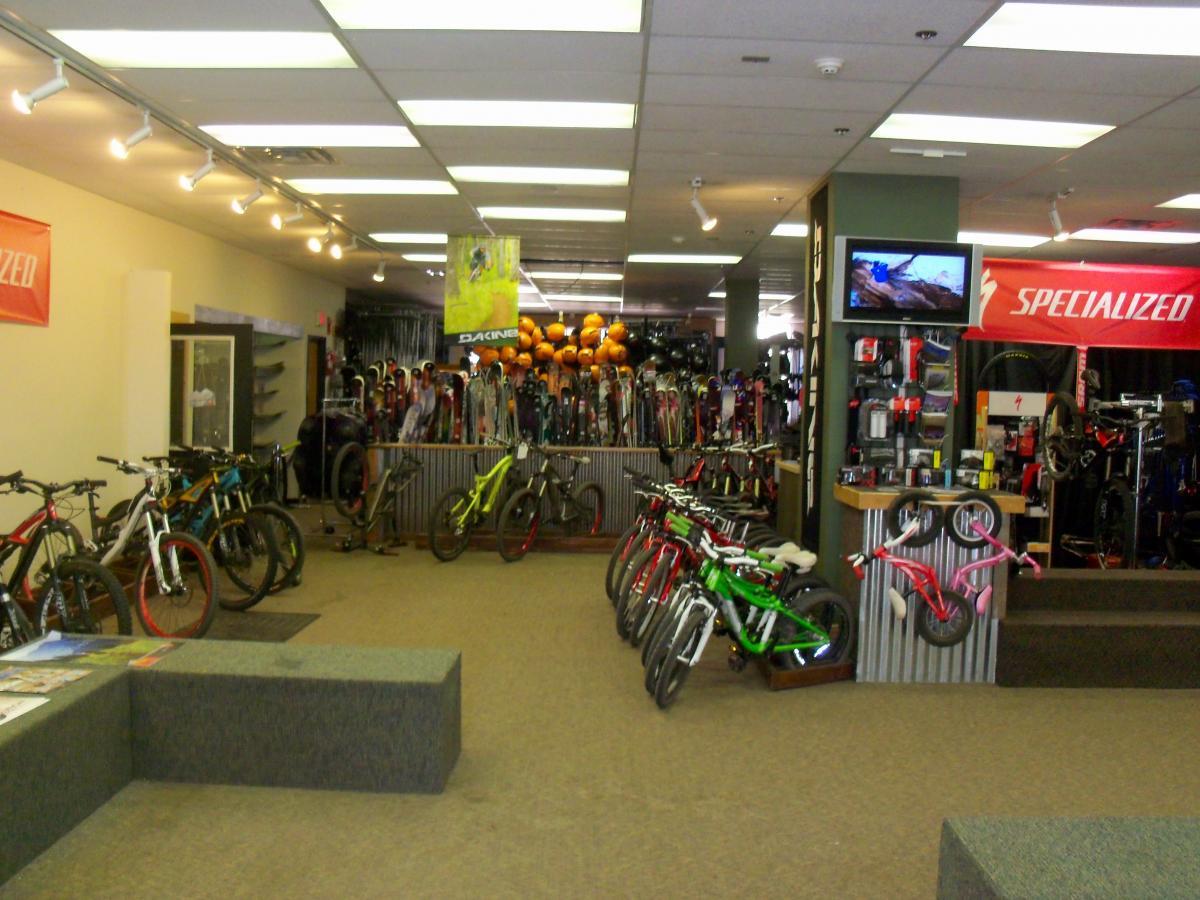 Interior of a sporting goods store featuring a variety of bicycles displayed prominently. In the background, there are shelves stocked with skiing gear and helmets. A television screen is visible, showing outdoor activity footage. The store has bright lighting and banners from the brands Specialized and Dakine. A seating area with brochures is located in the front. Evolution Bike Park at Crested Butte Mountain Resort mountain bike trail.