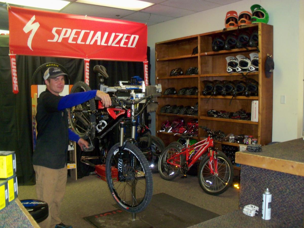 A bicycle mechanic working in a shop, holding a mountain bike wheel while standing next to a red bicycle. The background features various bicycle helmets and equipment on shelves, with a red "Specialized" banner displayed above. The shop has a clean, organized appearance, indicating a focus on bicycle maintenance and sales. Evolution Bike Park at Crested Butte Mountain Resort mountain bike trail.