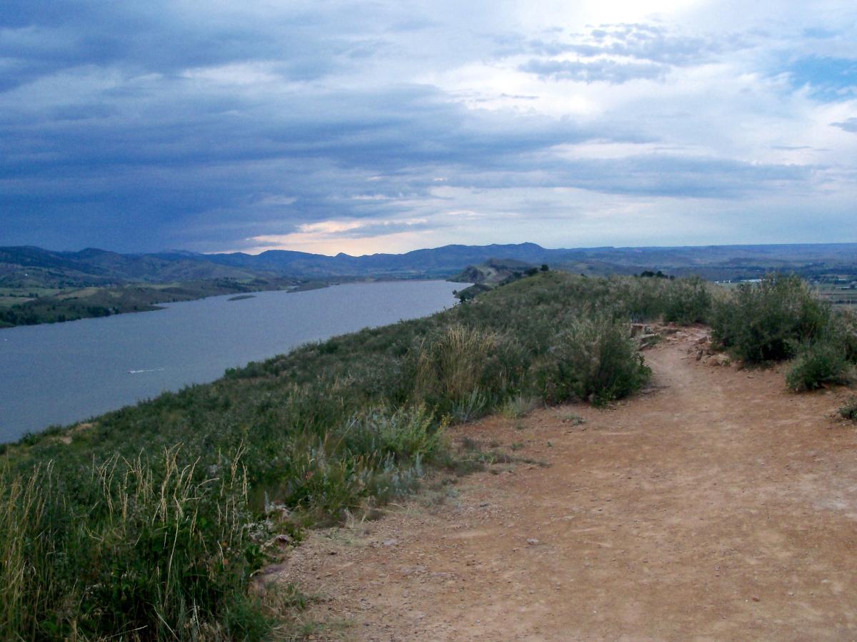 A scenic view overlooking a river surrounded by green hills and mountains under a dramatic sky with scattered clouds. A dirt pathway leads gently down toward the water's edge. Foothills Trail mountain bike trail.