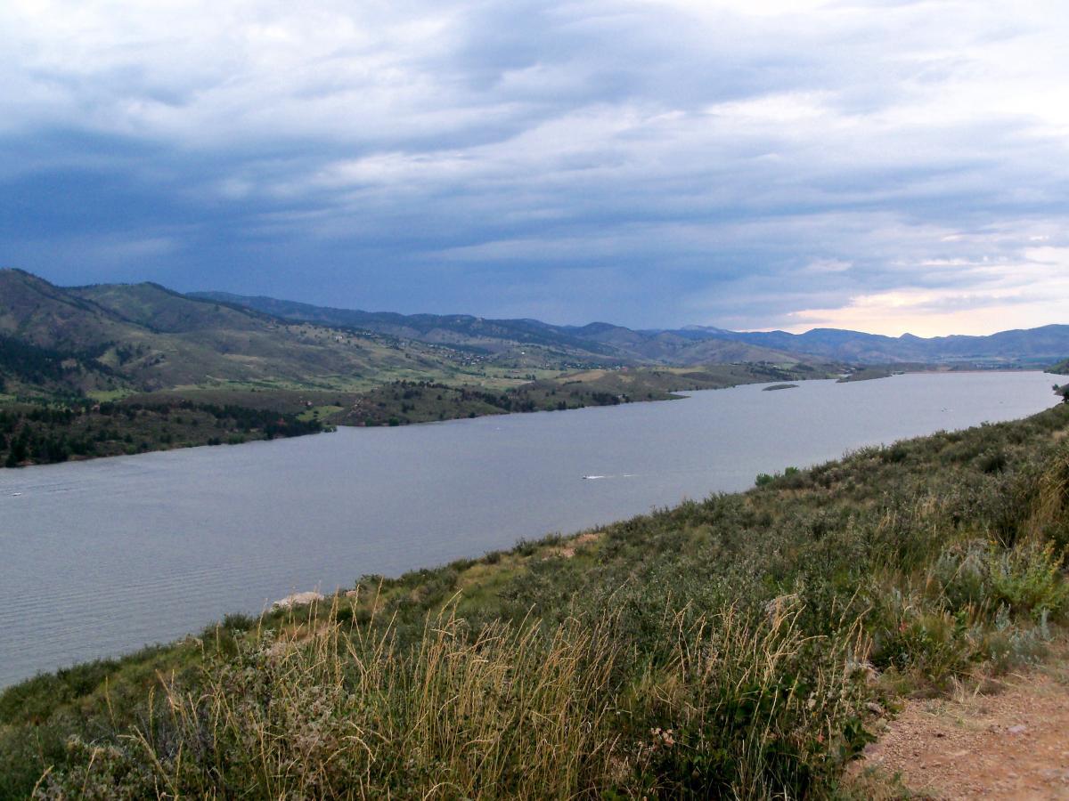 A panoramic view of a river winding through green hills under a cloudy sky, with patches of vegetation in the foreground. The scene captures the natural beauty of the landscape, highlighting the interplay of water, land, and clouds. Foothills Trail mountain bike trail.