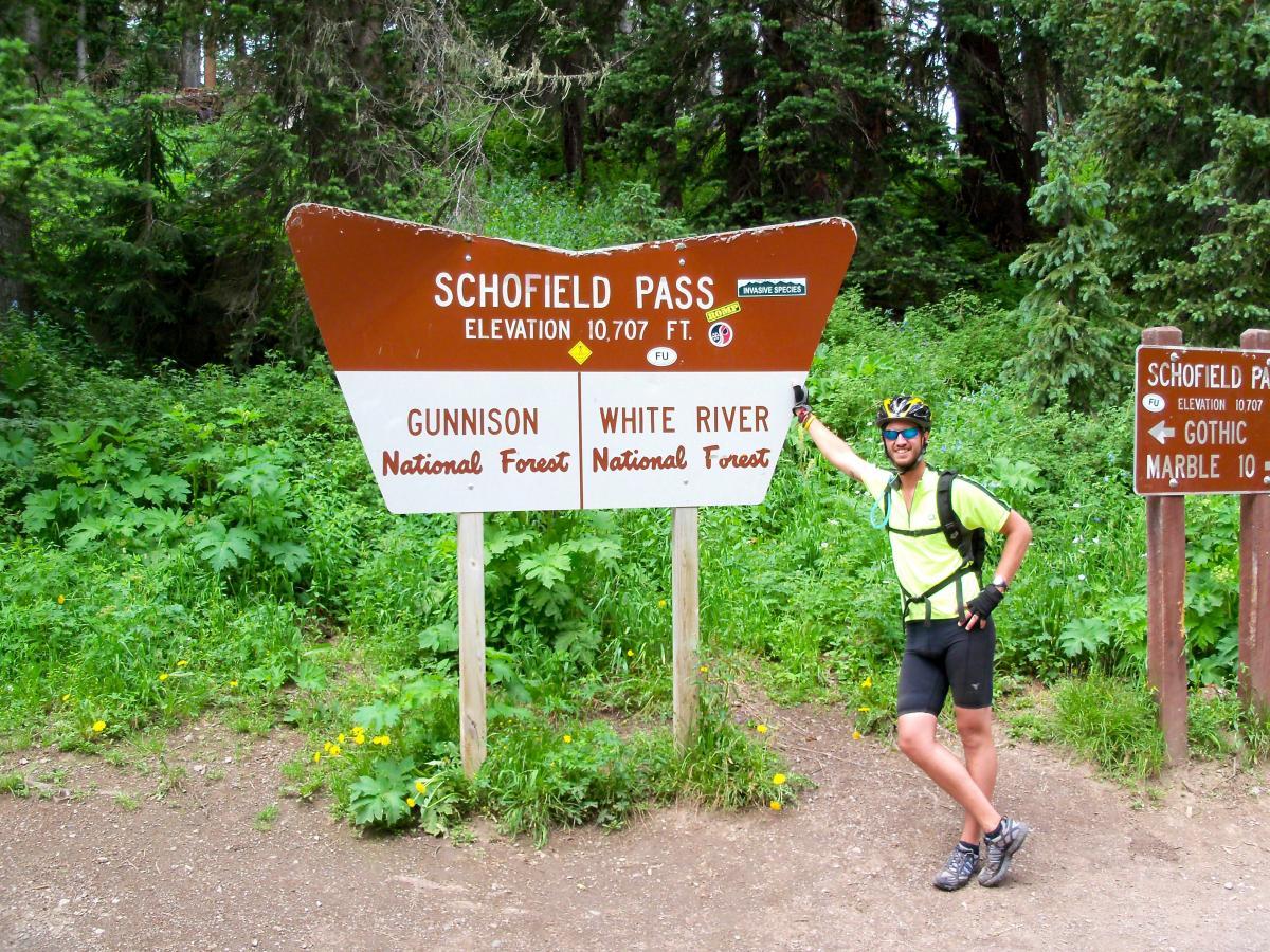 A cyclist stands beside a sign for Schofield Pass, which indicates an elevation of 10,707 feet. The sign is located in a lush forested area, with green vegetation and wildflowers surrounding it. The cyclist is wearing a bright yellow shirt and a helmet, with a friendly pose. Additional signage to the right provides directions to Gothic and Marble in the Gunnison and White River National Forests. Trail 401 mountain bike trail.