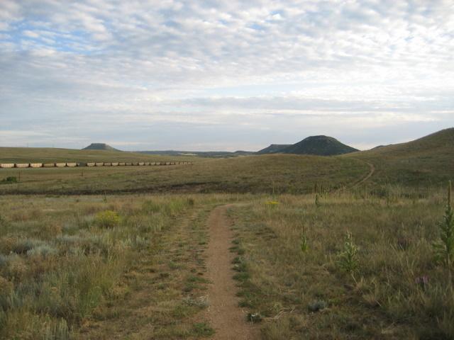 A wide, open landscape featuring rolling hills and a dirt path winding through grassy terrain. The sky is partly cloudy, and a faint outline of distant hills is visible. In the background, a line of train cars can be seen traveling along the flatlands. Greenland Open Space mountain bike trail.