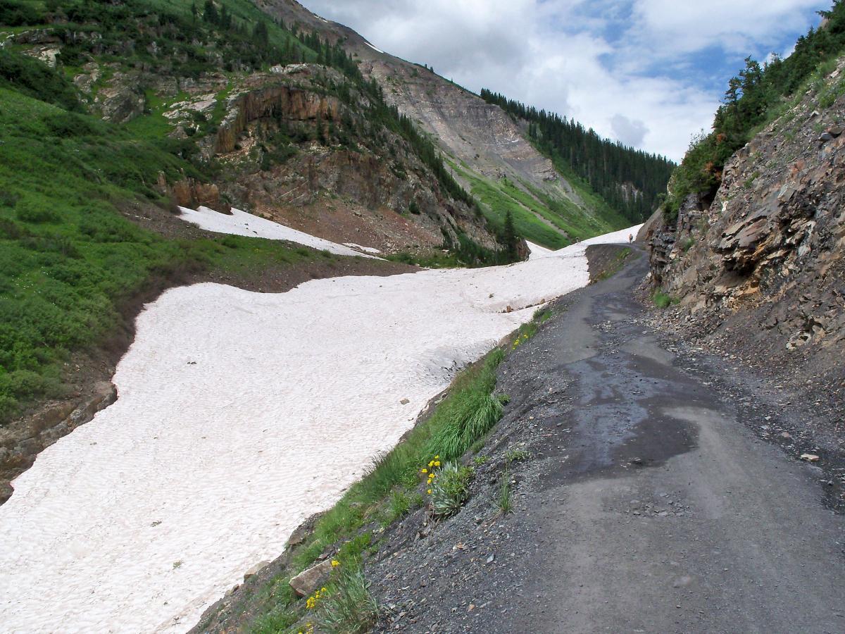 A winding dirt road alongside a snow patch in a mountainous area, with green vegetation on the slopes and rocky cliffs in the background under a partly cloudy sky. Trail 401 mountain bike trail.