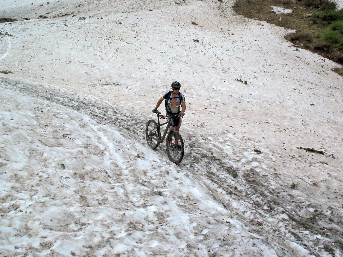 A mountain biker riding up a snowy slope, surrounded by patches of grass and rocky terrain. The cyclist is wearing a helmet and athletic clothing, with faint tire tracks visible in the snow. Trail 401 mountain bike trail.