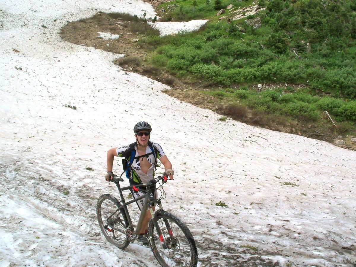 A person riding a mountain bike uphill on a snowy slope, surrounded by greenery. The cyclist is wearing a helmet and sunglasses, showcasing a determined expression. The background features a mixture of snow and patches of grass. Trail 401 mountain bike trail.