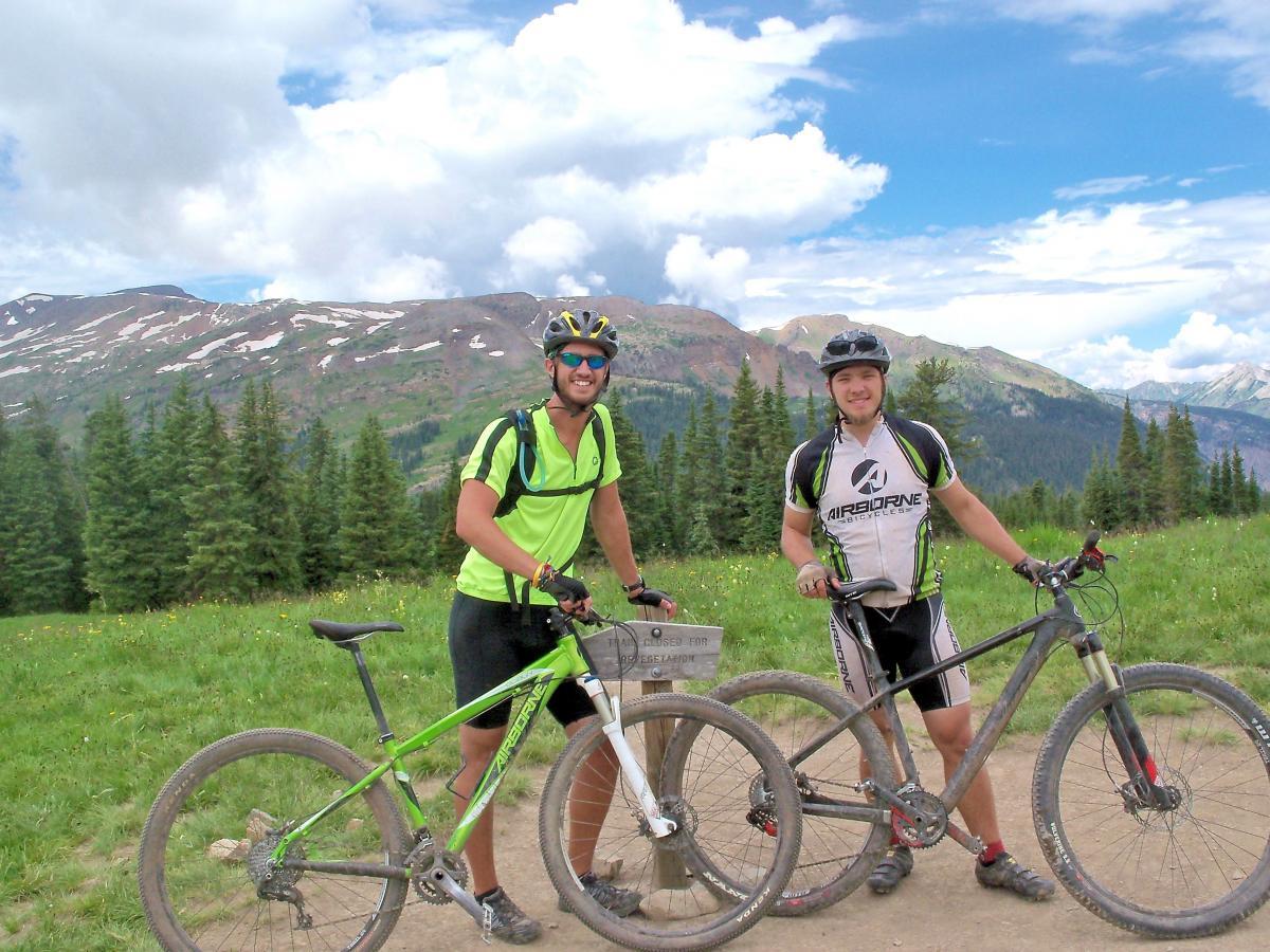 Two mountain bikers posing with their bikes on a scenic trail surrounded by green trees and mountains in the background. The sky is partly cloudy, and both riders are wearing helmets and athletic clothing. One biker is on the left wearing a bright yellow shirt, while the other is on the right in a black and white jersey. A wooden trail sign is visible nearby. Trail 401 mountain bike trail.