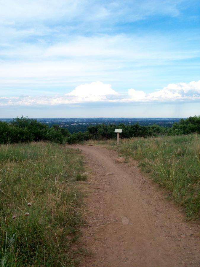 A dirt path winding through open grassland, leading towards a scenic view of distant hills and a vibrant blue sky filled with clouds. A small sign is visible along the path, indicating an informational point. Foothills Trail mountain bike trail.