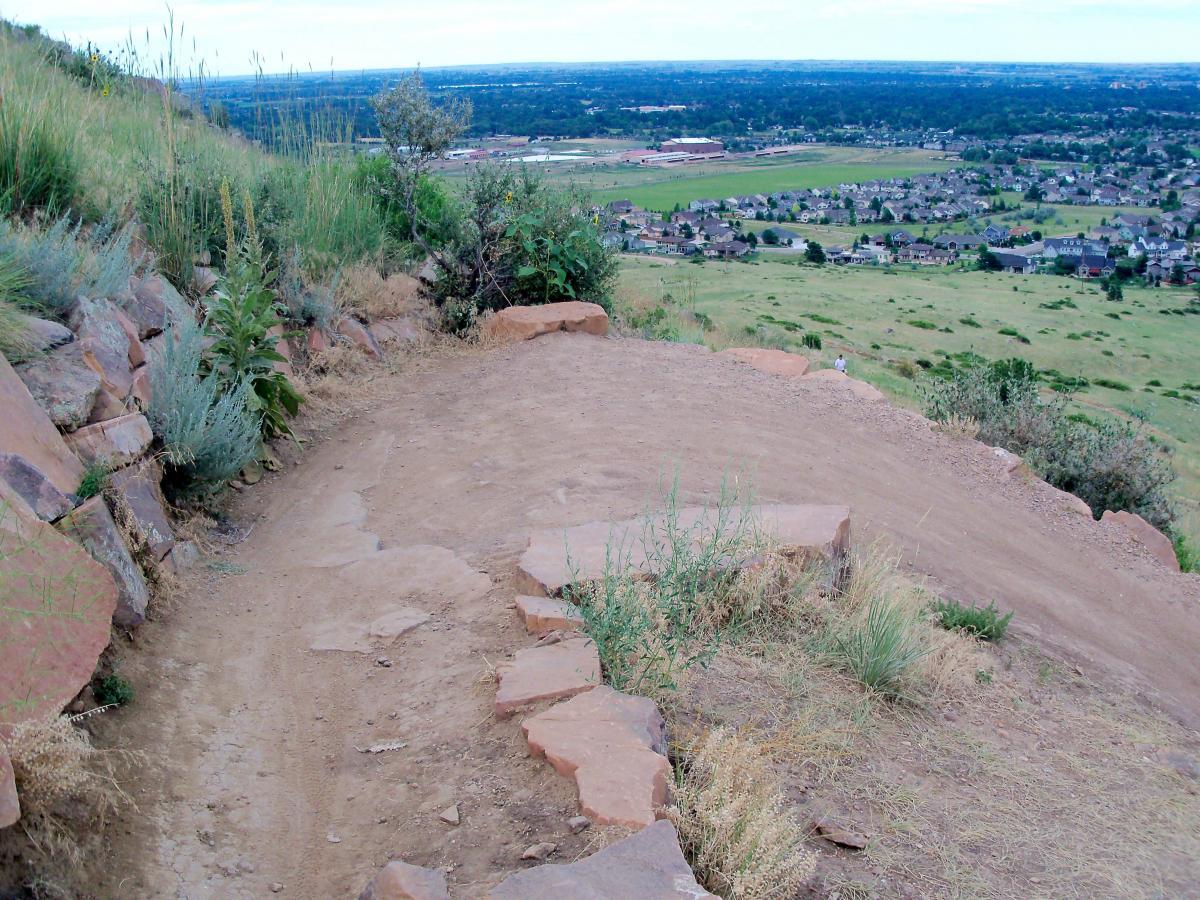 A rugged dirt path winds along the edge of a hillside, surrounded by sparse vegetation and rocky outcrops. In the background, a panoramic view reveals a sprawling landscape of fields and residential areas under a cloudy sky. Foothills Trail mountain bike trail.