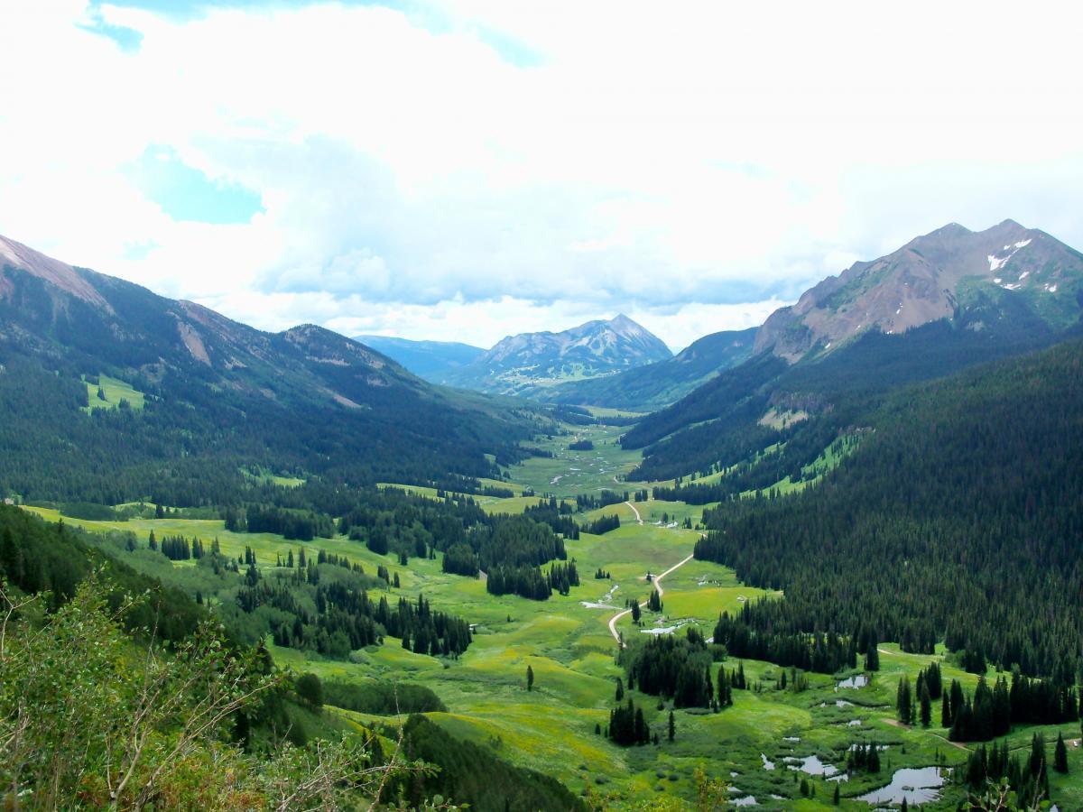 A scenic view of a lush green valley surrounded by mountains under a partly cloudy sky. The landscape features rolling hills, dense forests of evergreens, and winding roads leading through the vibrant meadow. Snow-capped peaks are visible in the background, enhancing the natural beauty of the area. Trail 401 mountain bike trail.