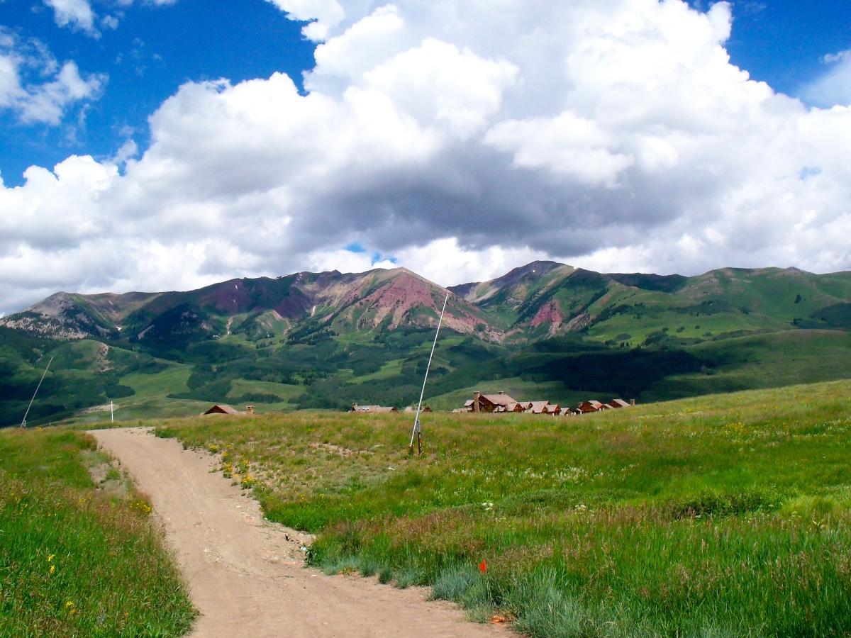 A scenic view of verdant hills and majestic mountains under a partly cloudy blue sky. A dirt path leads through a grassy field, with a few rustic buildings visible in the distance, framed by the vibrant greenery of summer. Evolution Bike Park at Crested Butte Mountain Resort mountain bike trail.