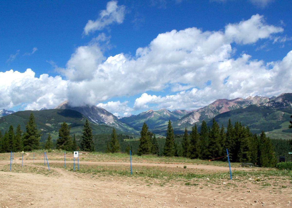 A scenic view of mountainous terrain featuring lush green hills and a variety of trees, set against a bright blue sky with fluffy white clouds. The foreground shows a dirt path bordered by blue poles, leading towards the expansive mountains in the background, some of which are partially covered by low-hanging clouds. Evolution Bike Park at Crested Butte Mountain Resort mountain bike trail.