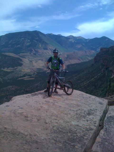 A mountain biker stands on a rock ledge, overlooking a vast landscape of valleys and mountains. The cyclist wears a helmet and a green and blue shirt and is positioned on a mountain bike. The backdrop features rolling hills and a partly cloudy sky. The Whole Enchilada mountain bike trail.