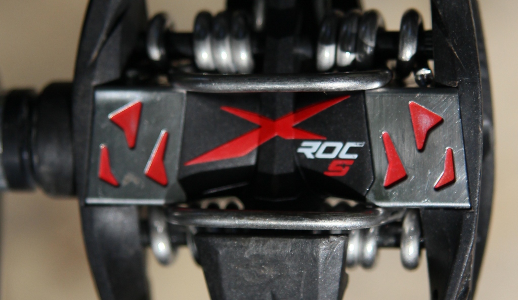 Close-up view of a black bicycle pedal featuring red accents and the logo "XROC S" prominently displayed in the center. The design includes several metallic springs and structural components, emphasizing its functional and sporty aesthetic.
