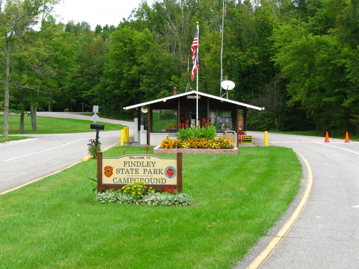 Image of the entrance to Findley State Park Campground, featuring a welcome sign, colorful flower beds, a small building with a roof, and trees in the background. A winding road leads towards the entrance, with traffic cones placed nearby. Findley State Park mountain bike trail.