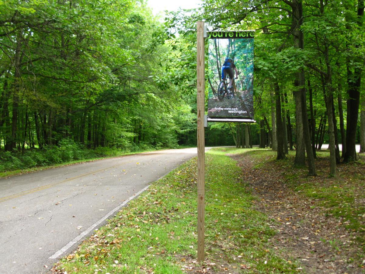 A forested area alongside a road featuring a signpost displaying a map and the text "you're here." The sign depicts a mountain biker on a trail, indicating the location as Findley State Park in Wellington. Green trees and foliage surround the scene, creating a natural, outdoor atmosphere. Findley State Park mountain bike trail.