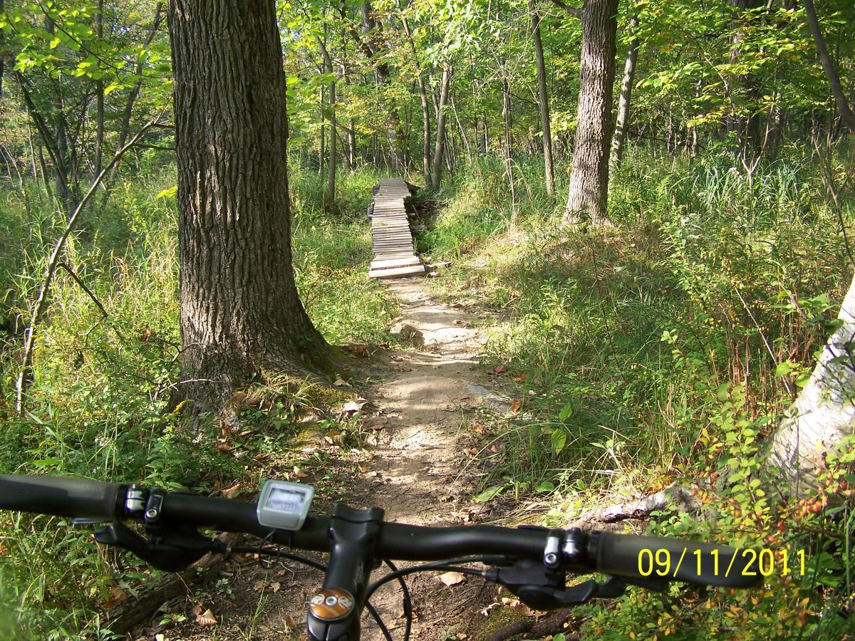 View from a mountain bike handlebars along a forest trail, showcasing a wooden bridge crossing a grassy area. Surrounding trees and underbrush create a lush green environment. The date in the corner reads September 11, 2011. Edwards Creek mountain bike trail.