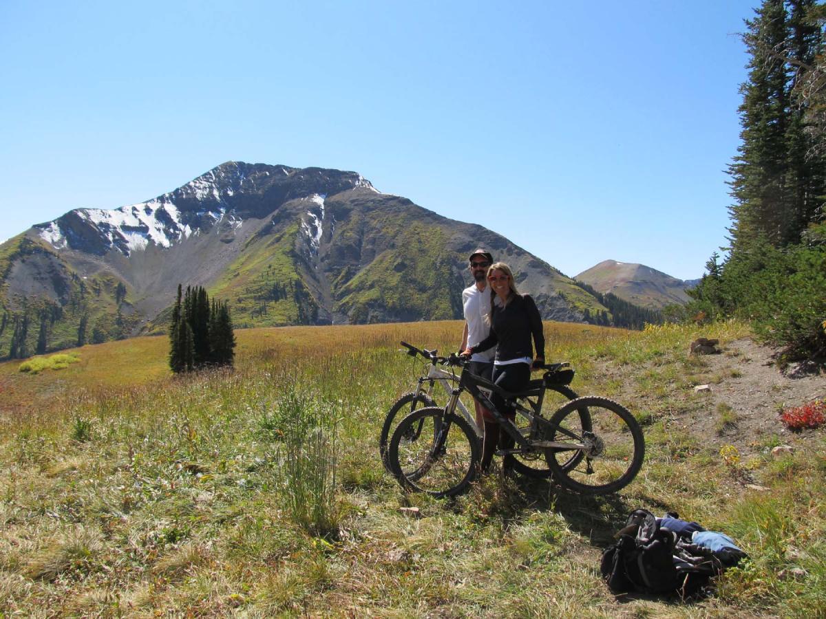 A couple standing with mountain bikes in a grassy field, with a scenic, snow-capped mountain backdrop under a clear blue sky. Trail 401 mountain bike trail.