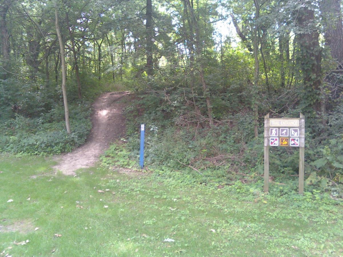 A dirt path leads into a wooded area, flanked by lush green grass and foliage. A sign labeled "Trail Entrance" with various icons indicating trail rules and restrictions is visible on the right side. The path has a gentle incline, suggesting a natural transition into the forest. Espenscheid Forest Perserve mountain bike trail.
