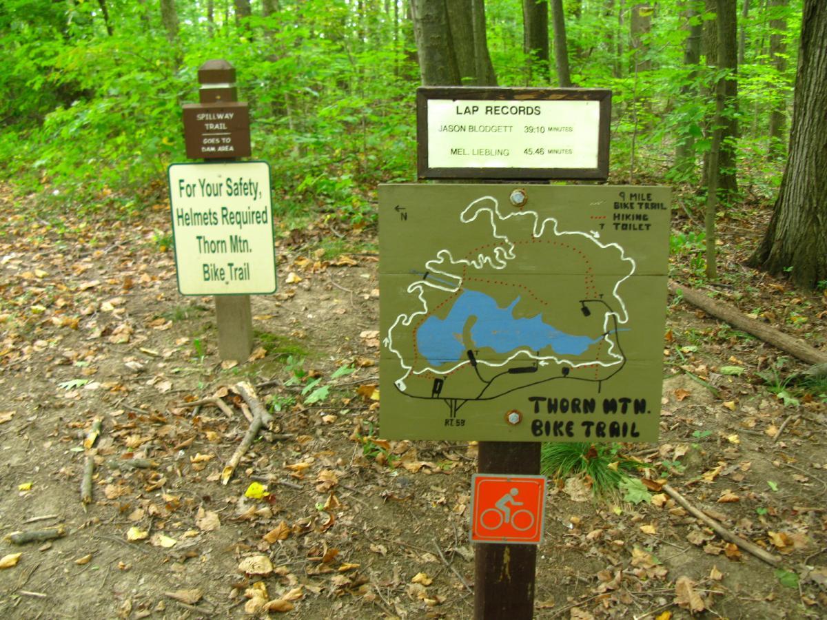 A sign at a forested trailhead featuring a detailed map of the Thorn Mountain Bike Trail, indicating a 9-mile bike trail with hiking options and restrooms. The sign includes lap records for two individuals and a warning about mandatory helmet use for safety. The area is surrounded by lush green trees and scattered leaves on the ground. Findley State Park mountain bike trail.