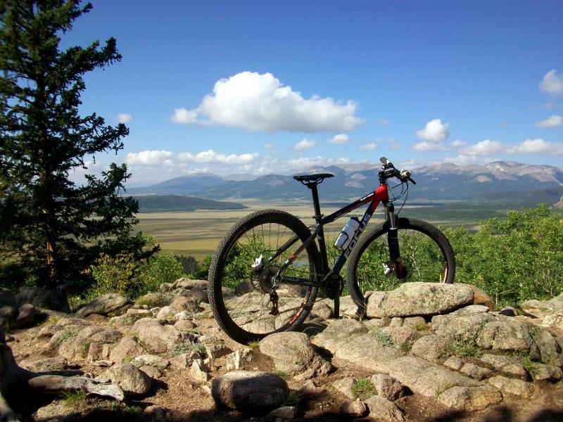 A mountain bike resting on a rocky outcrop, with a scenic view of rolling hills and mountains in the background. The sky is largely clear with a few fluffy clouds scattered across it. Surrounding vegetation includes trees and greenery, indicating a natural outdoor setting. Colorado Trail: Kenosha Pass To Breckenridge mountain bike trail.