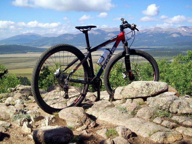 A mountain bike resting on rocks with a scenic view of mountains and a valley in the background under a partly cloudy sky. Colorado Trail: Kenosha Pass To Breckenridge mountain bike trail.