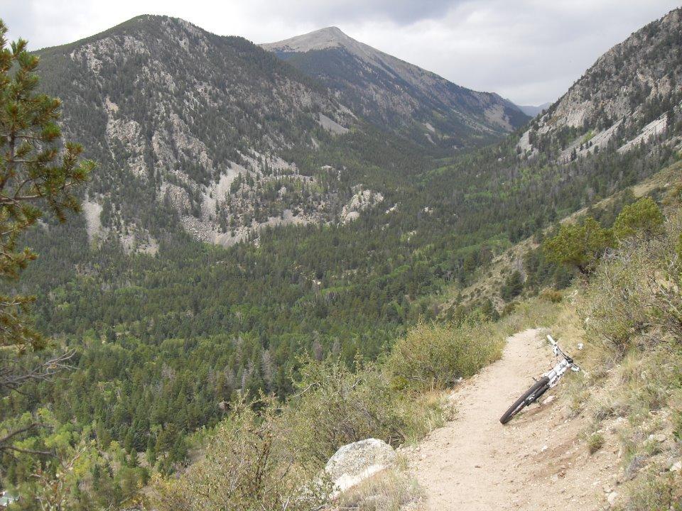 A scenic view of a mountainous landscape with a lush green valley below, featuring a dirt bike trail winding through the bushes. A mountain bike lies on its side on the trail, with rolling hills and rocky peaks in the background under a cloudy sky. Colorado Trail mountain bike trail.