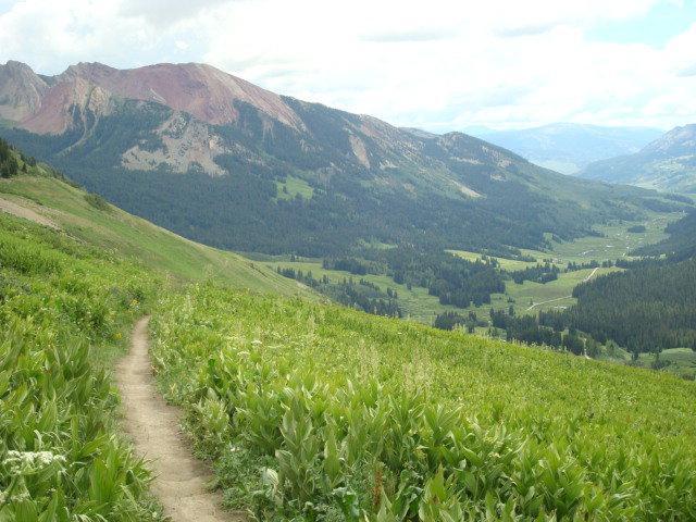 A scenic view of a winding dirt path leading through lush green meadows, set against a backdrop of majestic mountains with varying shades of green and reddish-brown. The sky is partly cloudy, adding depth to the landscape. Trail 401 mountain bike trail.
