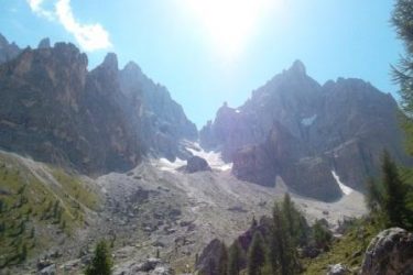 A scenic view of towering rocky mountains against a clear blue sky, with sunlight peeking over the peaks. Lush green trees line the foreground, and a glacier is visible in the mountain's valley. Val Venegia - Dolomites mountain bike trail.