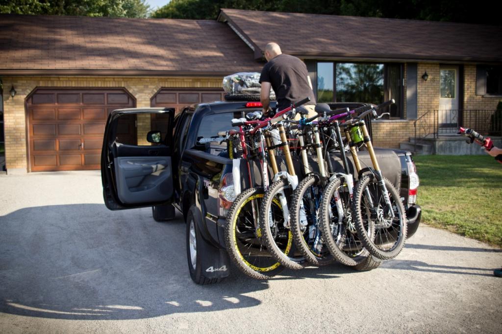 A person loading several mountain bikes onto the back of a black pickup truck parked in a driveway. The truck has its back open, and some bikes are already secured in place. The house in the background features a brown roof and two garage doors. The scene is set in a sunny outdoor environment. Seven Springs mountain bike trail.