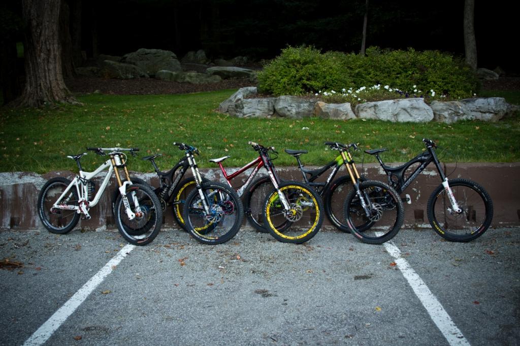 A row of six mountain bikes lined up in a parking area, with a background of grass and rocks. The bikes feature various colors and designs, showcasing their unique styles and features. Seven Springs mountain bike trail.