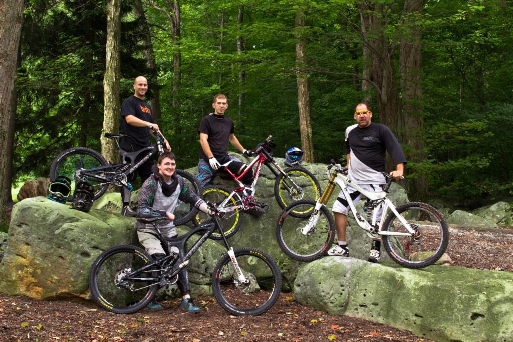 A group of four mountain bikers posing on large rocks in a forested area. Each rider is standing with their bike, showcasing different styles and colors. The background features lush green trees, creating a vibrant outdoor setting. Seven Springs mountain bike trail.