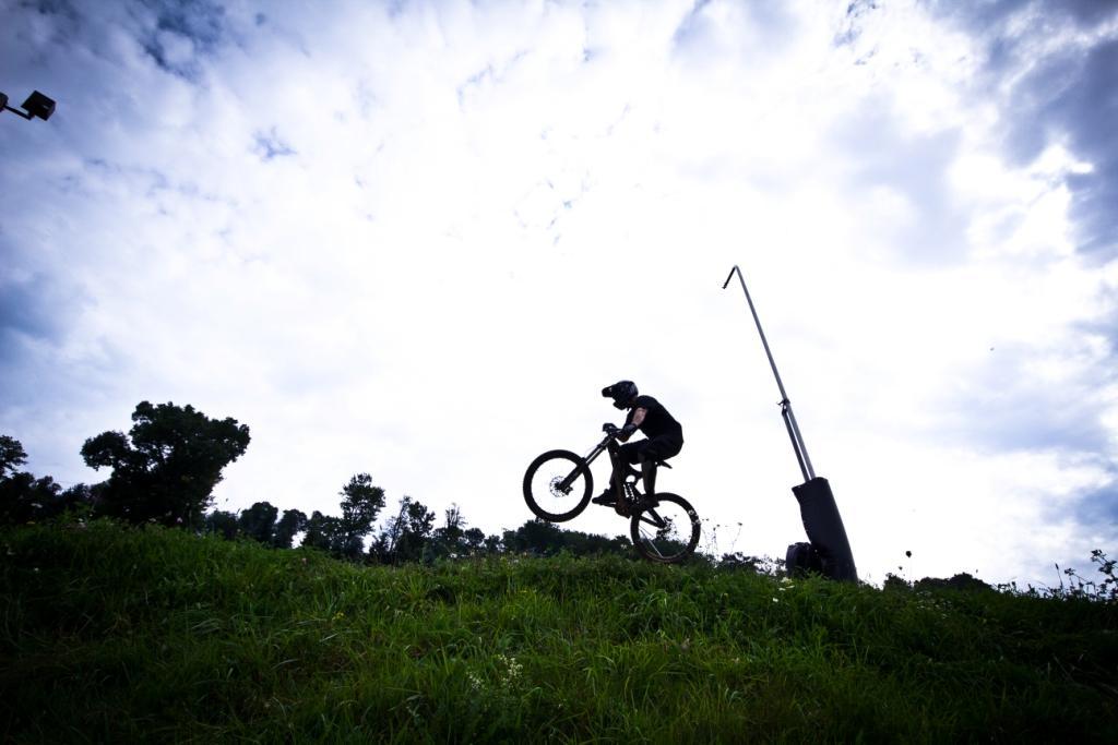 A silhouette of a mountain biker in mid-air, performing a jump over a grassy hill, with dark clouds in the sky and trees in the background. Seven Springs mountain bike trail.