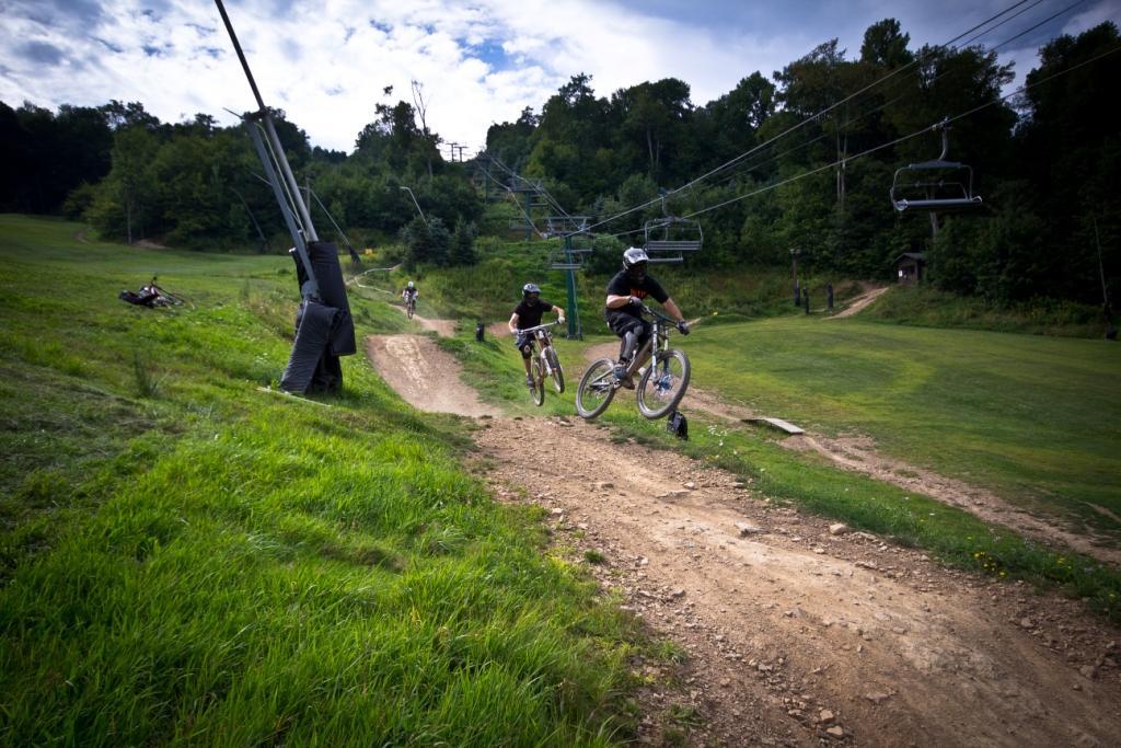 A group of mountain bikers riding on a dirt trail, with one cyclist airborne over a jump. In the background, a chairlift ascends a hill surrounded by trees. The landscape features green grass and a clear sky. Seven Springs mountain bike trail.