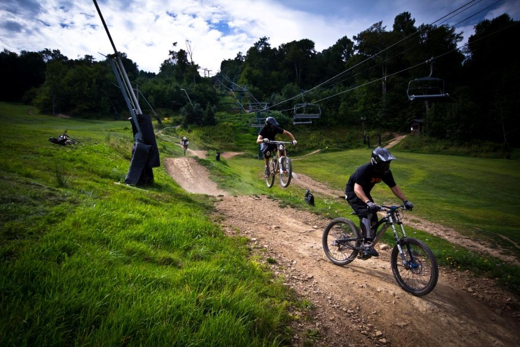 A group of mountain bikers riding down a dirt path in a green valley, with one individual airborne while jumping off a ramp. Chairlifts are visible in the background, and the scene is set against a backdrop of trees and a partly cloudy sky. Seven Springs mountain bike trail.