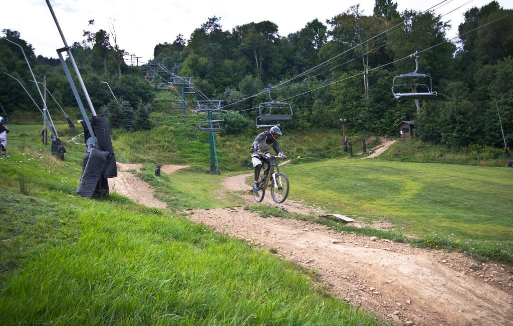 A mountain biker riding on a dirt trail in a green hillside area, with ski lift chairs visible in the background, surrounded by trees and grass. Seven Springs mountain bike trail.