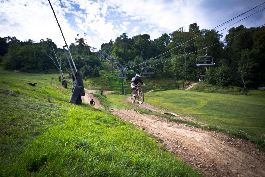 A mountain biker in a helmet and protective gear jumps off a dirt ramp while riding downhill on a grassy slope. In the background, a ski lift with empty chairs runs alongside the trail, surrounded by trees under a partly cloudy sky. Seven Springs mountain bike trail.