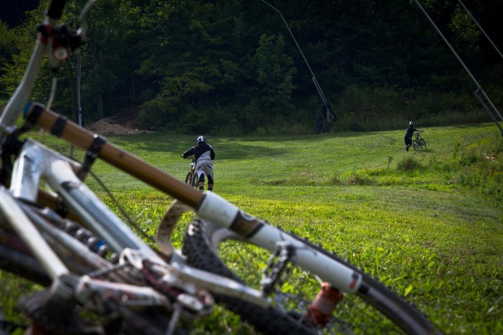 A scenic view of a grassy hill with two mountain bikers riding in the background, while several bicycles are lying in the foreground. The scene is set in a forested area with trees visible in the backdrop, indicating an outdoor biking park environment. Seven Springs mountain bike trail.