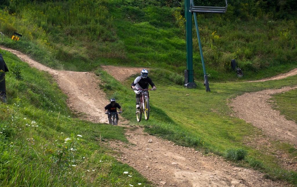 Two mountain bikers navigate a dirt trail in a green landscape, with one rider airborne as he jumps off a small ramp while the other rides closely behind on the ground. In the background, a ski lift is visible, surrounded by lush vegetation. Seven Springs mountain bike trail.
