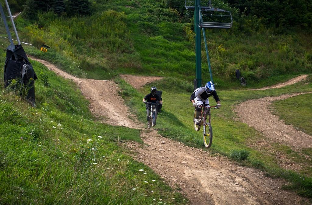 Two mountain bikers navigate a dirt trail in a grassy area, with one rider airborne while the other follows closely. In the background, a ski lift can be seen. The scene captures a vibrant outdoor setting suitable for biking, surrounded by greenery and a winding path. Seven Springs mountain bike trail.