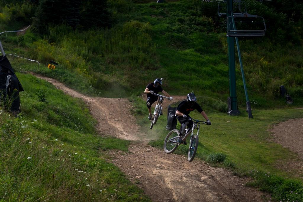 Two mountain bikers performing tricks on a dirt path surrounded by lush greenery. One biker is airborne, while the other is navigating a turn on the ground. In the background, a ski lift is visible, indicating a hilly terrain suitable for biking. Dust is kicked up from the trail, showcasing the action and excitement of the sport. Seven Springs mountain bike trail.