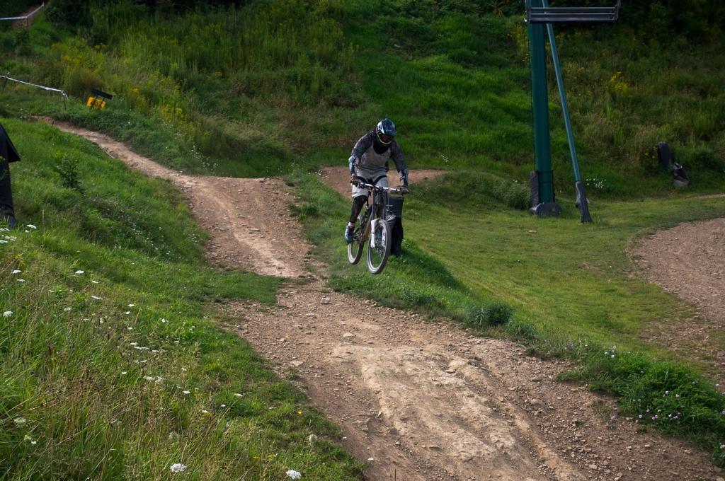 A mountain biker performing a jump on a dirt track in a grassy landscape, with a ski lift visible in the background. Seven Springs mountain bike trail.