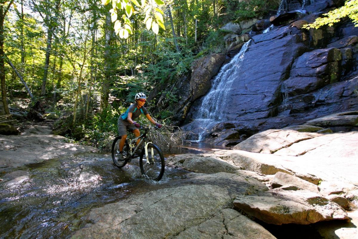 A mountain biker riding through a rocky area near a waterfall, surrounded by lush green trees and natural scenery, with water splashing around the bike. Vallee Bras Du Nord Secteur Shannahan mountain bike trail.