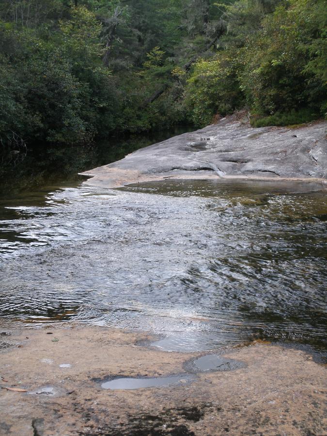 A serene landscape featuring a rocky riverbank with smooth stones and gentle water flow. Lush greenery surrounds the scene, providing a tranquil natural setting. Panthertown Valley mountain bike trail.