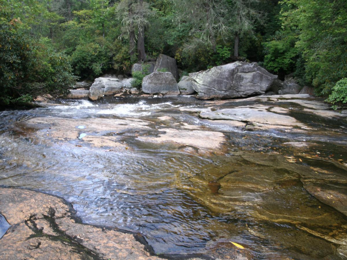 A serene view of a gently flowing river surrounded by lush greenery and large rocks. The water reflects the surrounding environment, with visible ripples and smooth rocky surfaces. Panthertown Valley mountain bike trail.