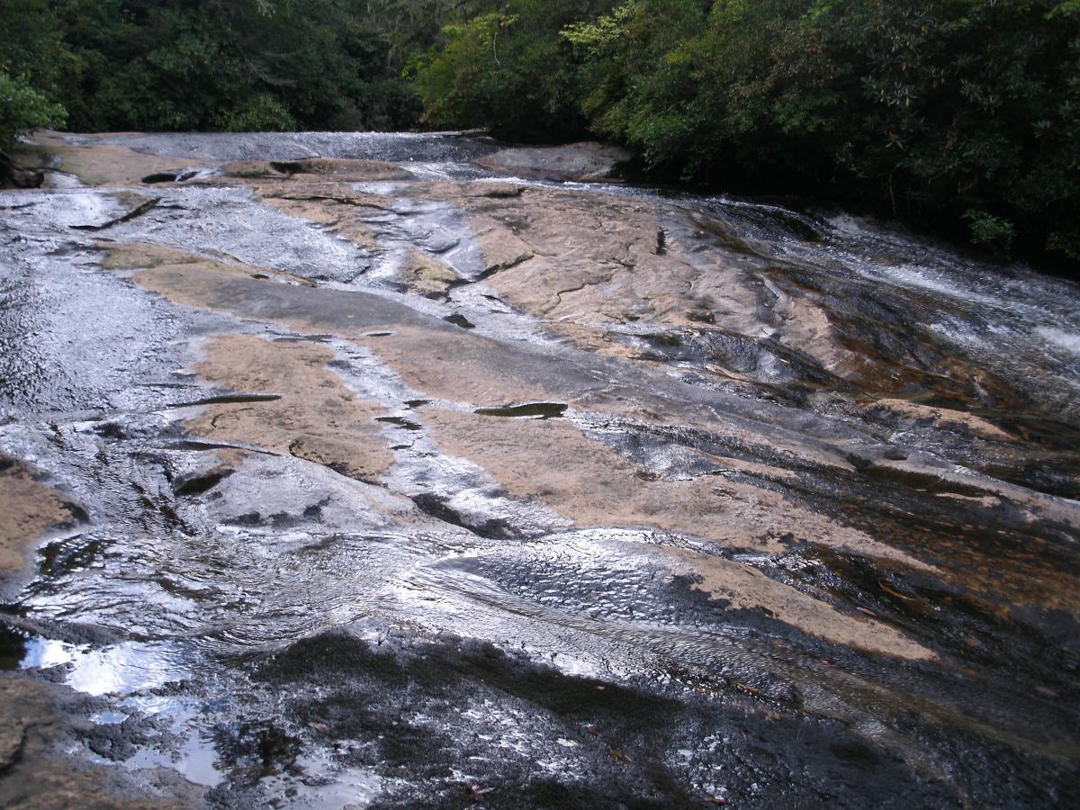A smooth, rocky surface of a waterfall or riverbed, with water flowing over it, surrounded by lush green foliage. The contrasting colors of wet and dry rock create a natural, serene landscape. Panthertown Valley mountain bike trail.
