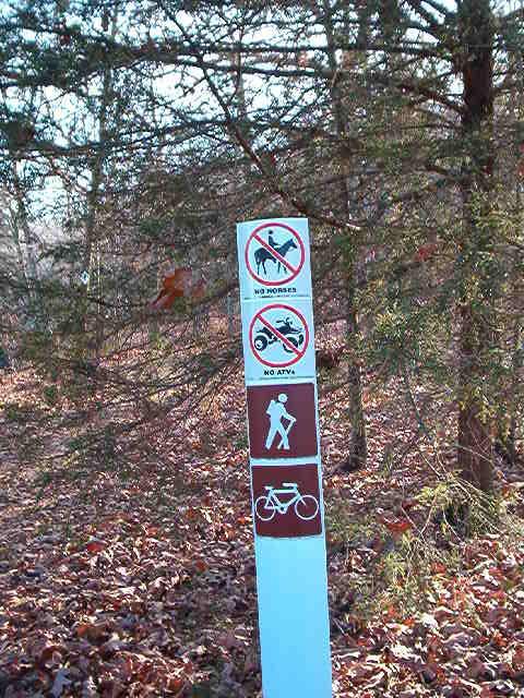 A trail sign surrounded by trees, displaying multiple symbols: a "no horses" sign, a "no ATVs" sign, and a sign indicating that hiking and biking are allowed. The ground is covered in fallen leaves. Lincoln Lake mountain bike trail.